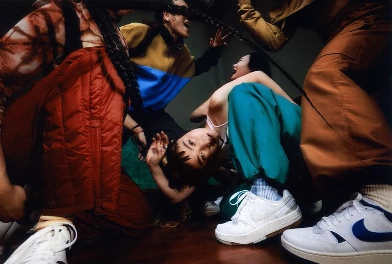Multiple people gathered indoors, showcasing a variety of colorful clothing and footwear, including white Nike sneakers, in an ensemble shot.