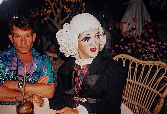 Two people sit at a table outdoors. One is a man in a patterned shirt. The other is a person in drag with heavy makeup and a dark suit.