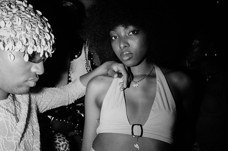 Two young adults pose indoors in a close-up studio shot. One wears a seashell headpiece and sunglasses, the other a halter top.