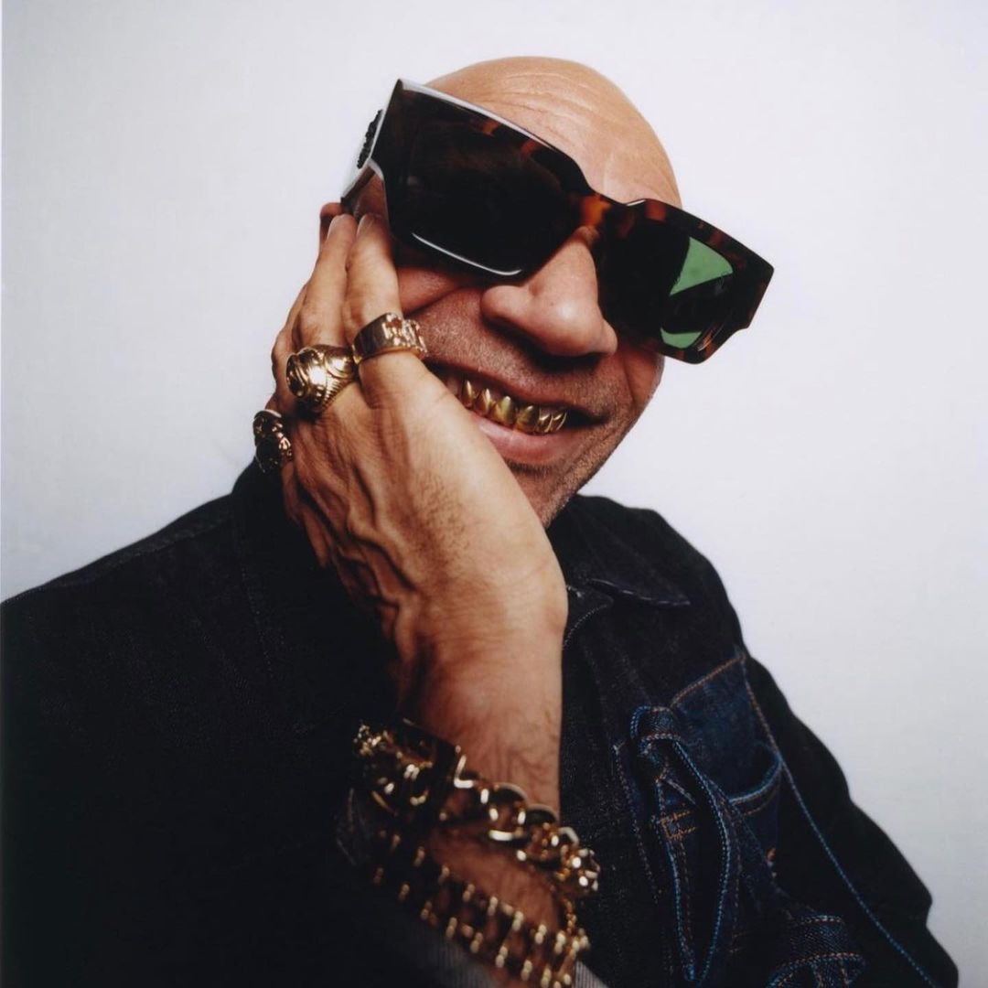 Close-up studio shot of a bald adult man with gold teeth and sunglasses, wearing a denim jacket and gold jewelry against a white background.