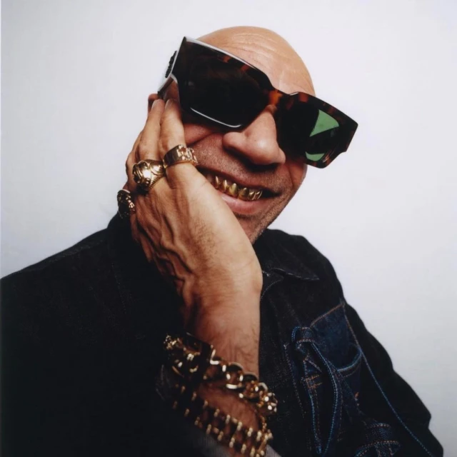 Close-up studio shot of a bald adult man with gold teeth and sunglasses, wearing a denim jacket and gold jewelry against a white background.