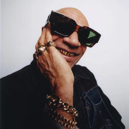 Close-up studio shot of a bald adult man with gold teeth and sunglasses, wearing a denim jacket and gold jewelry against a white background.