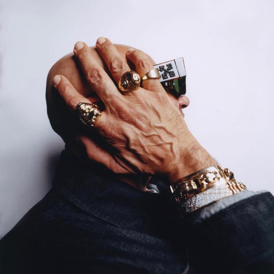 Close up of a bald adult male wearing sunglasses, gold rings, and bracelets against a light grey background in a studio setting.