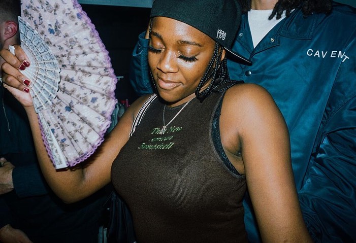 Young woman with braided hair and baseball cap in club, holding floral hand fan, wearing brown top with white straps and silver necklace.