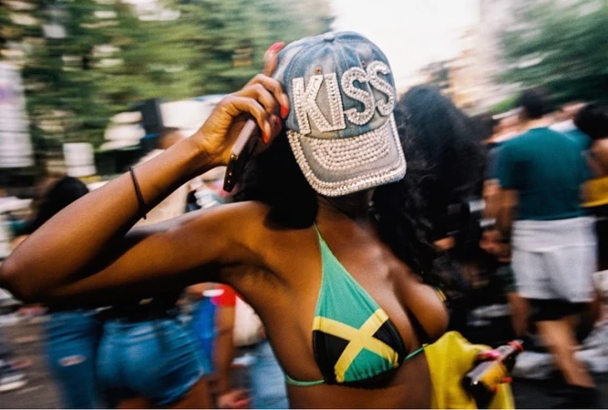 Woman in Jamaican flag bikini and bedazzled 'KISS' cap in a blurry outdoor crowd scene with cell phone, creating a vibrant, lively atmosphere.