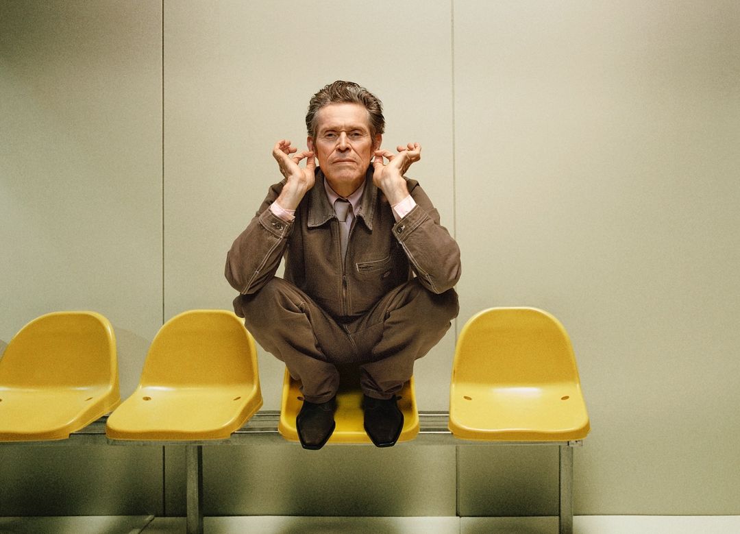 Willem Dafoe sits cross-legged on a yellow chair in a waiting room, holding his hands to his ears, looking directly at the camera.