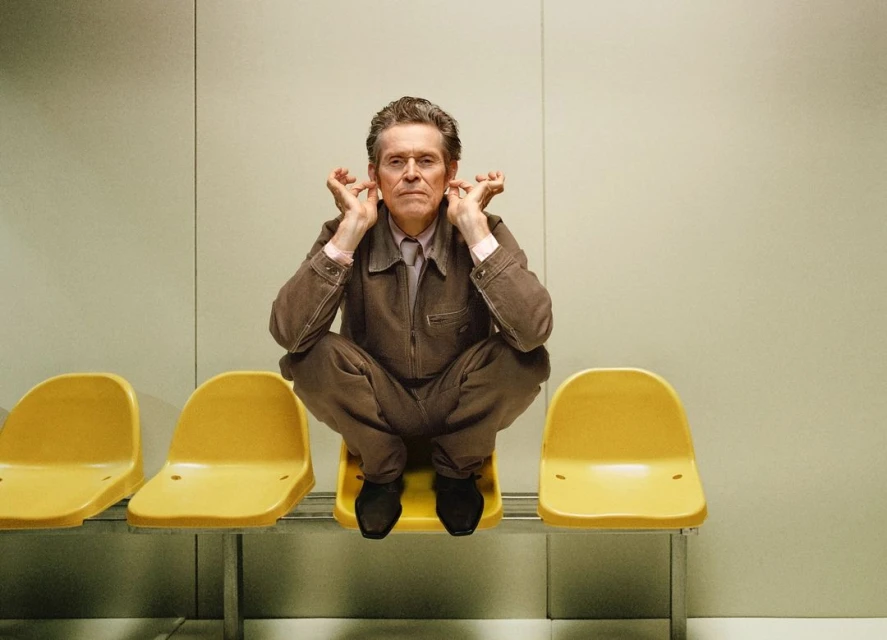 Willem Dafoe sits cross-legged on a yellow chair in a waiting room, holding his hands to his ears, looking directly at the camera.