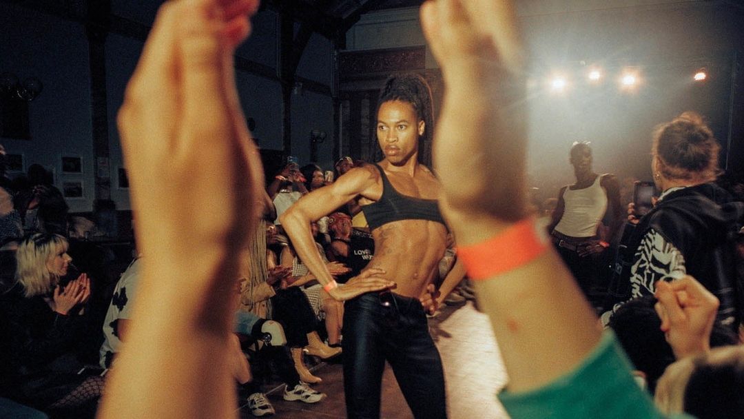Runway model poses during fashion show as crowd watches and applauds, illuminated by bright stage lights in a large, indoor venue.