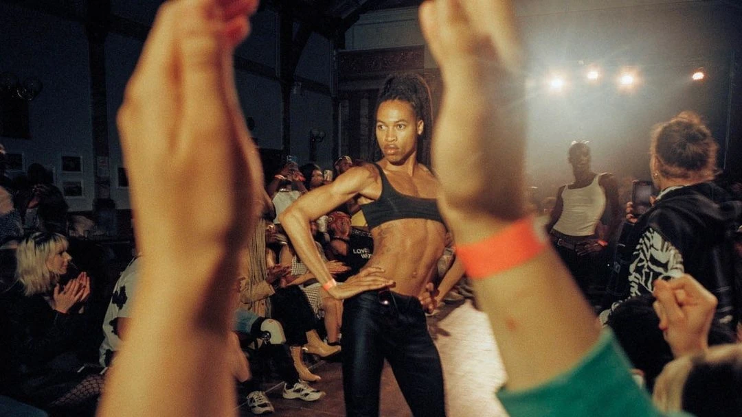 Runway model poses during fashion show as crowd watches and applauds, illuminated by bright stage lights in a large, indoor venue.