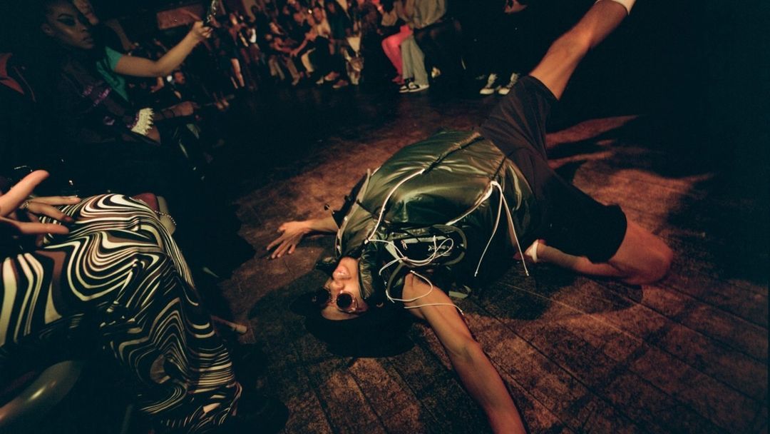 Low-angle shot of a man breakdancing on a wood floor in a dark room with a crowd of people in the background.