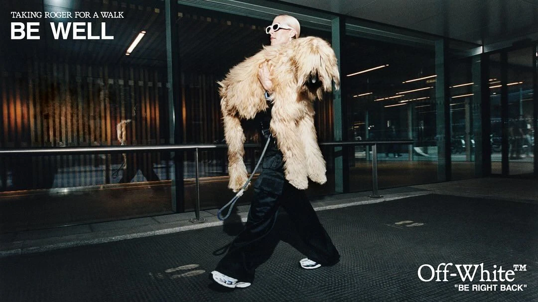 Person carrying a large fluffy dog-like object outside a building with glass windows, wearing sunglasses and a black outfit.