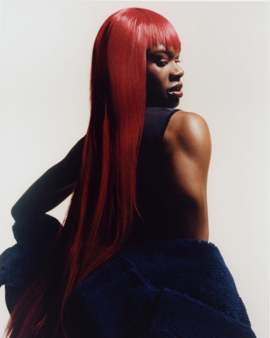 Portrait of a woman with long, bright red hair, wearing a dark top and navy blue jacket, posing in a studio setting with soft lighting.