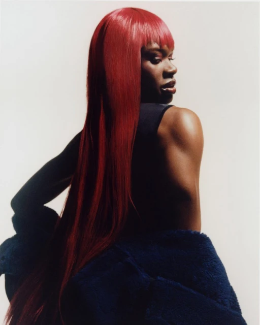 Portrait of a woman with long, bright red hair, wearing a dark top and navy blue jacket, posing in a studio setting with soft lighting.
