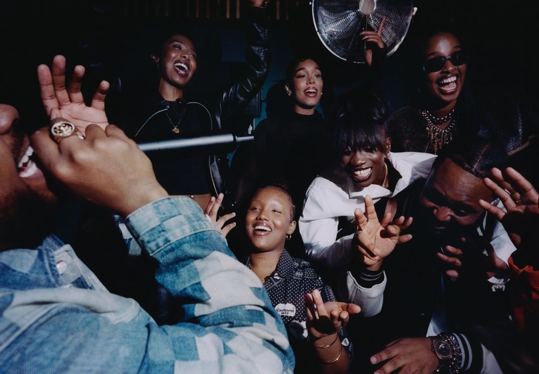 Group of diverse young adults laughing and singing in a dark indoor setting with a fan in the background, creating a lively atmosphere.