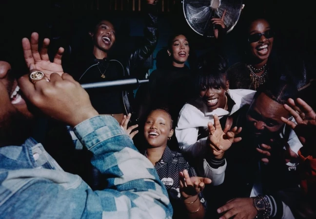 Group of diverse young adults laughing and singing in a dark indoor setting with a fan in the background, creating a lively atmosphere.