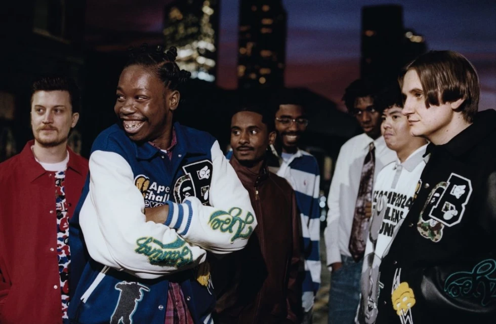 Group of young adult men pose together outdoors at dusk in a medium shot featuring varsity jackets and casual fashion in an urban setting.