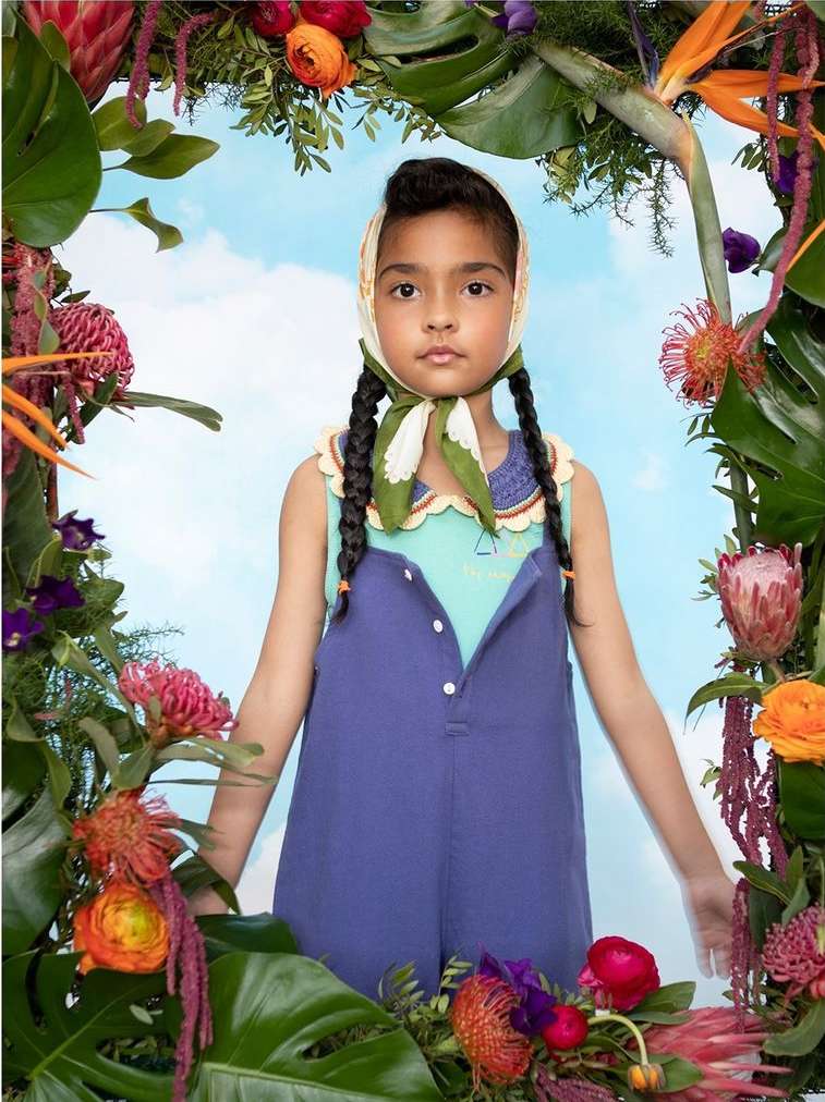 Portrait of a girl framed by colorful tropical flowers and foliage. She wears a blue jumpsuit and headscarf against a sky background.