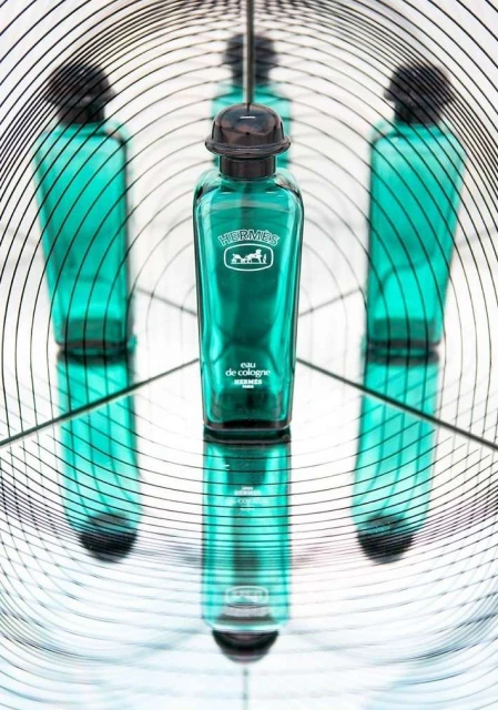 Hermes Eau de Cologne bottle stands reflecting in mirrors, surrounded by abstract geometric lines in an indoor setting, with a minimalist aesthetic.