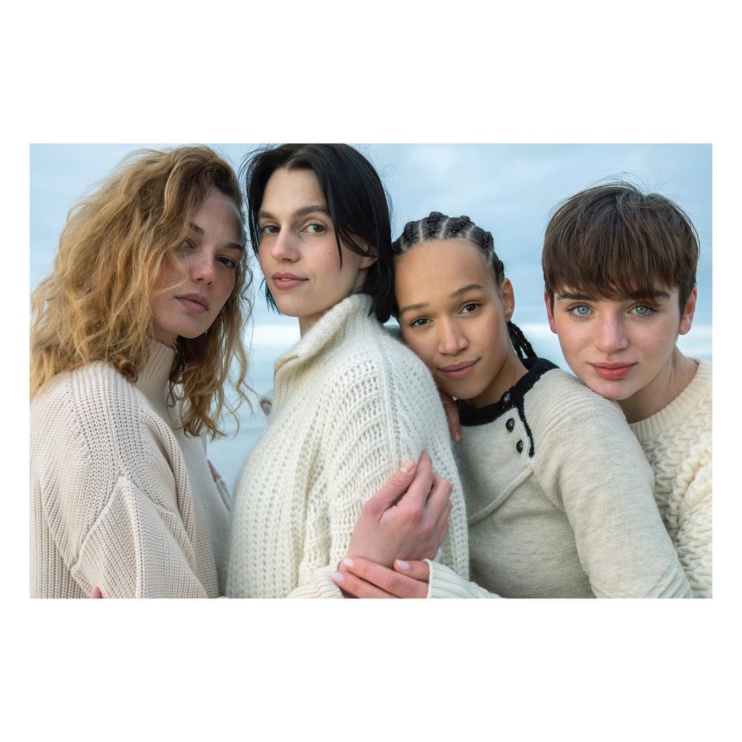 Four women in white sweaters pose together in front of a neutral sky, creating a portrait of friendship and coordinated fashion.