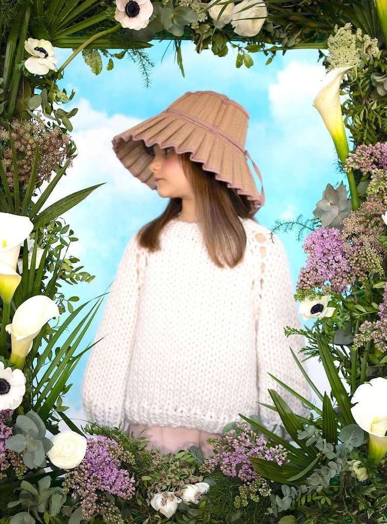 Young girl wears a wide-brimmed hat and cream sweater surrounded by flowers against a blue sky with clouds in a portrait.