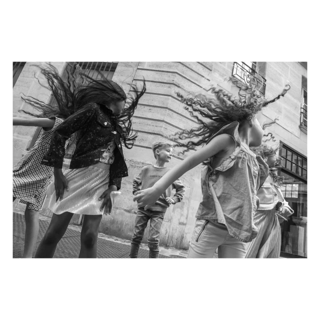 Group of children in motion, hair flying, wearing casual clothing, and standing outdoors against a building in a black and white photograph.