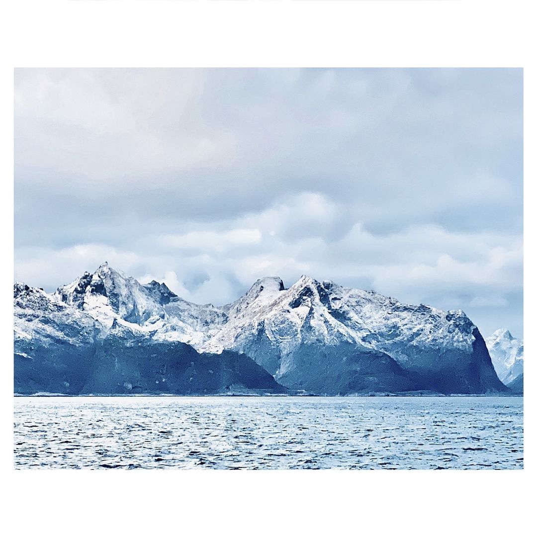 Scenic view of snow-covered mountains rising from the ocean under a cloudy sky, showcasing a serene winter landscape.