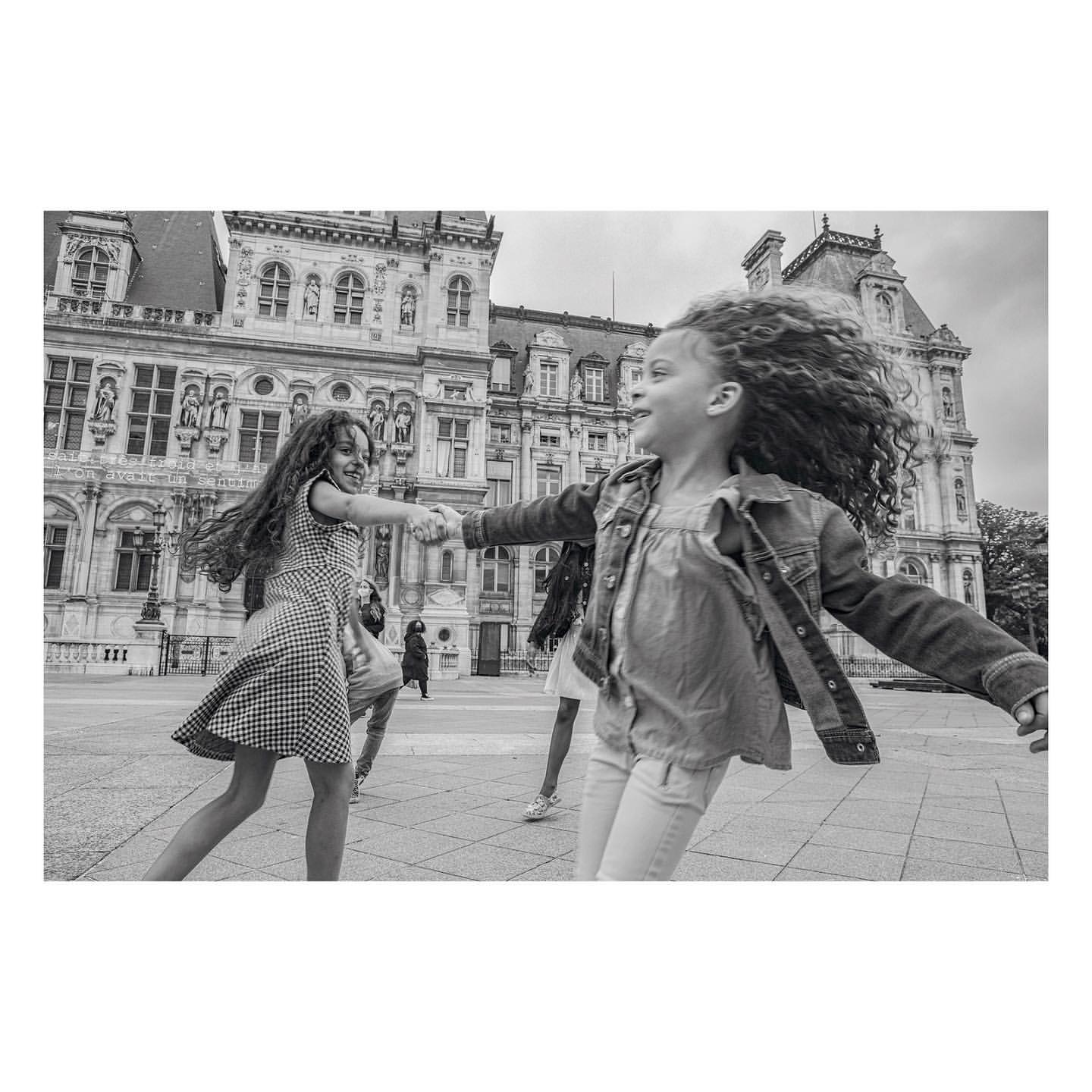 Two young girls in dresses, holding hands and spinning around in front of a large, ornate building on a black and white photo.