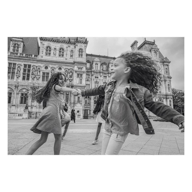 Two young girls in dresses, holding hands and spinning around in front of a large, ornate building on a black and white photo.