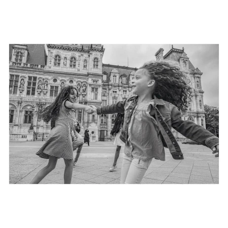 Two young girls in dresses, holding hands and spinning around in front of a large, ornate building on a black and white photo.