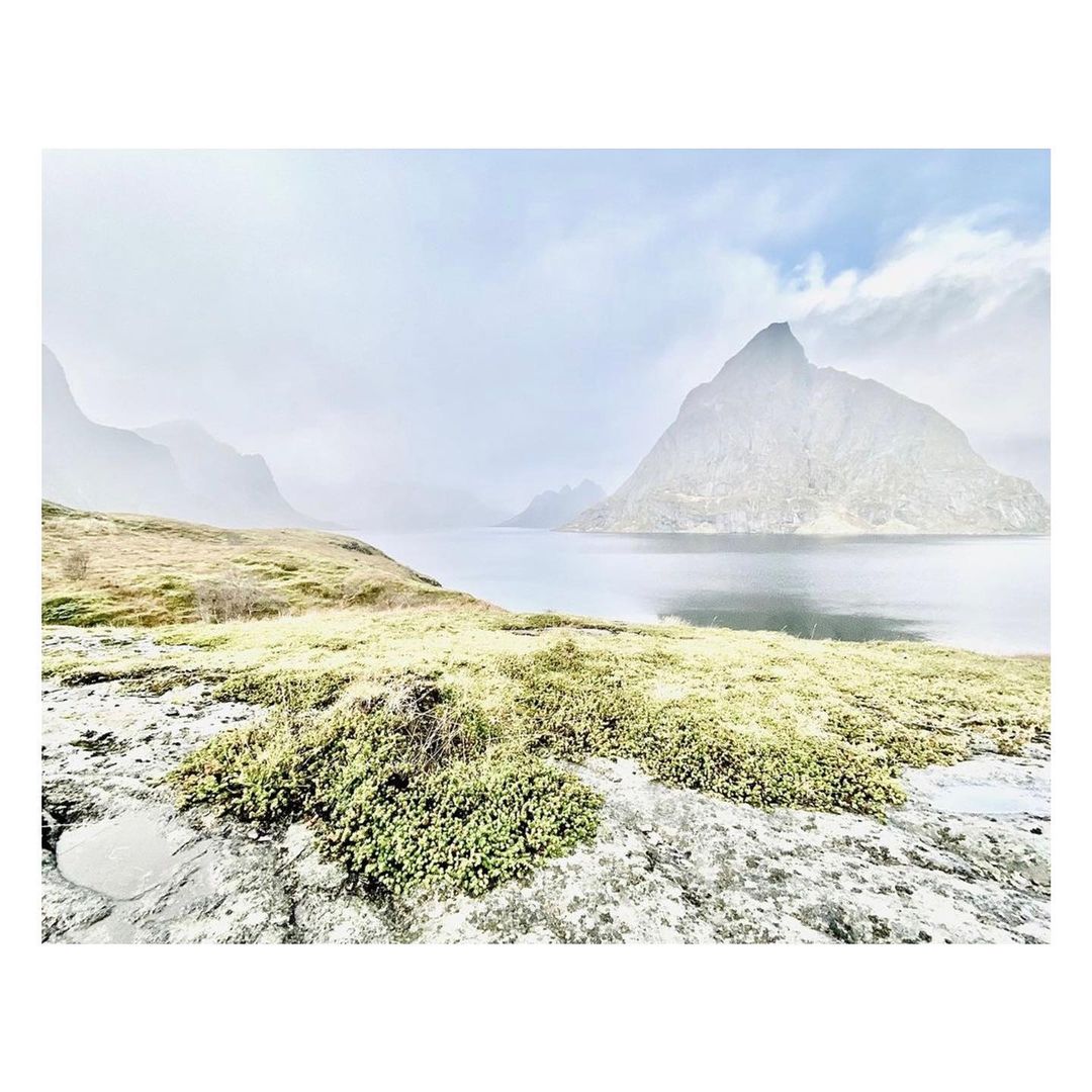 Scenic landscape featuring a tranquil body of water surrounded by grassy terrain, rugged mountains, and a cloudy sky, creating a serene atmosphere.
