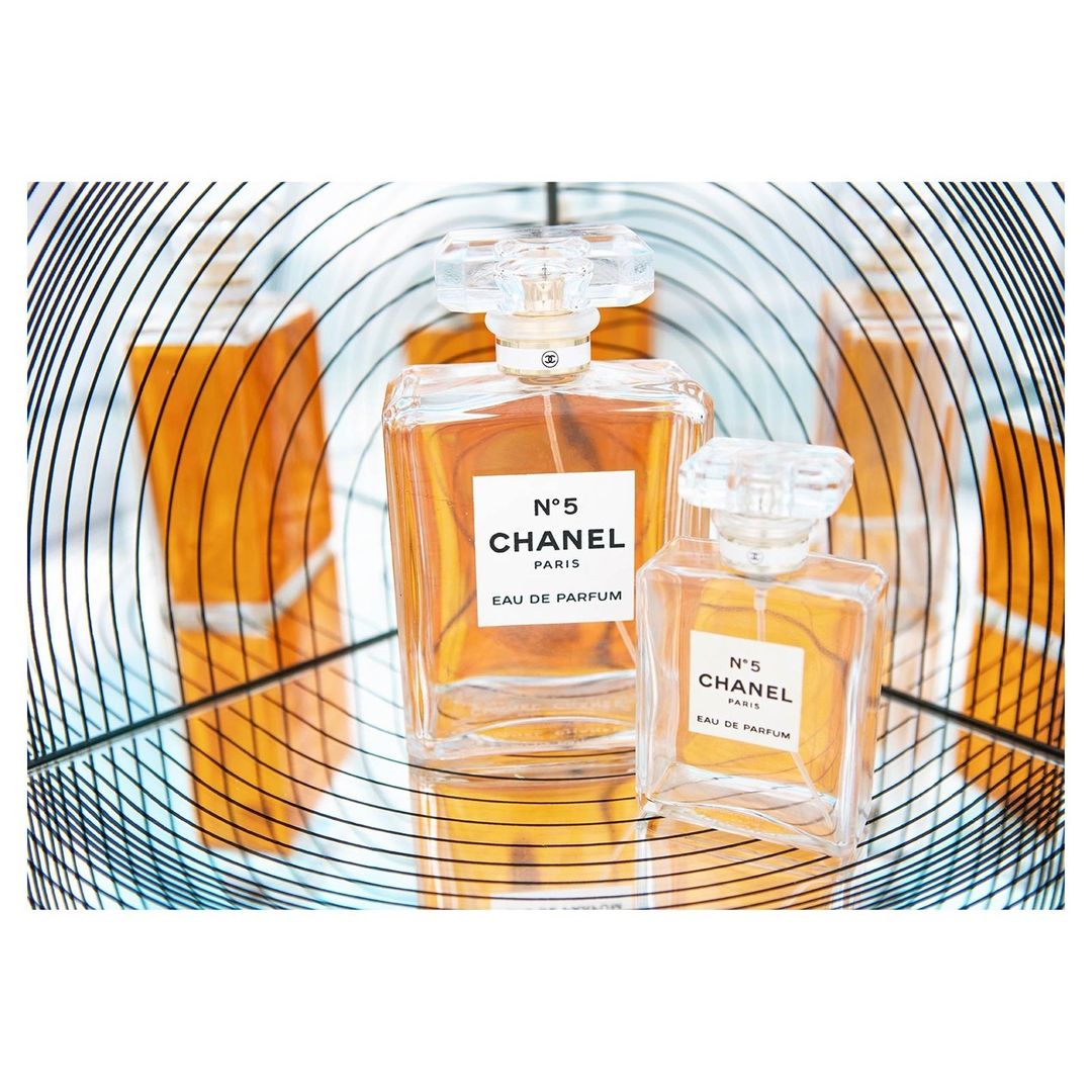 Close-up studio shot of two Chanel No. 5 perfume bottles of different sizes, set against a reflected amber and spiral background.