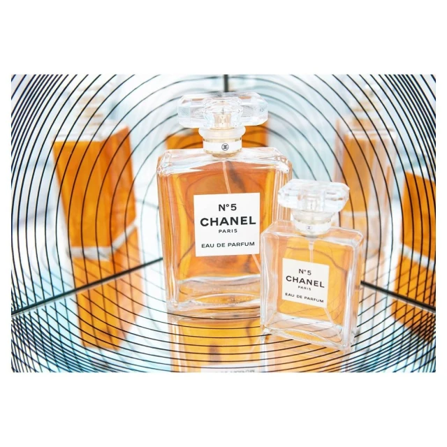 Close-up studio shot of two Chanel No. 5 perfume bottles of different sizes, set against a reflected amber and spiral background.