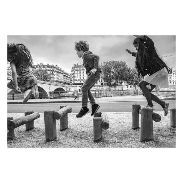 Three children jump from a wooden play structure outdoors, with a cityscape, bridge, and trees in the background. Black and white photo.