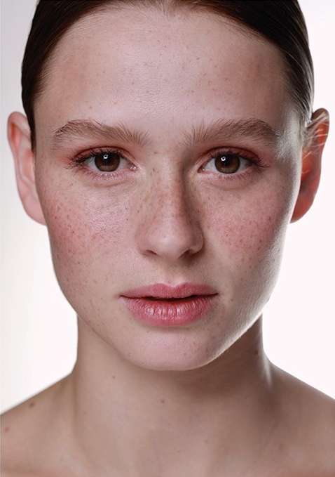 Close-up portrait of a young adult female with brown hair and freckles, looking directly at the camera against a bright white backdrop.