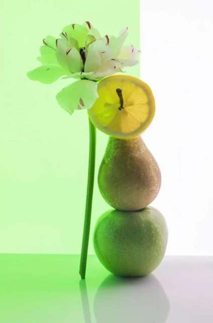 Still life of stacked green apple, pear, lemon slice, and white tulip against a split green and white background in a studio.