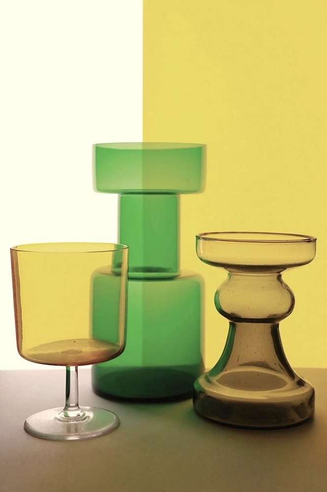Still life studio shot of a yellow wine glass, a green glass vase, and a smaller tan glass vase against a split yellow/white background.