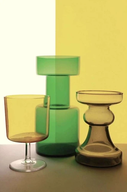 Still life studio shot of a yellow wine glass, a green glass vase, and a smaller tan glass vase against a split yellow/white background.