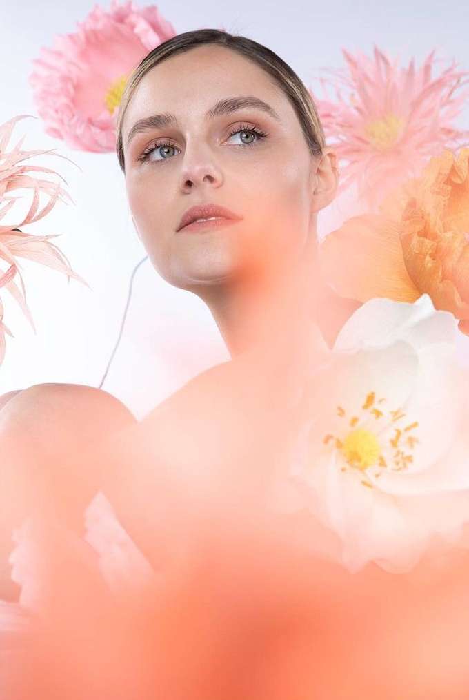 Portrait of a woman surrounded by pink, white, and peach flowers, posing in front of a white background in a beauty-style image.