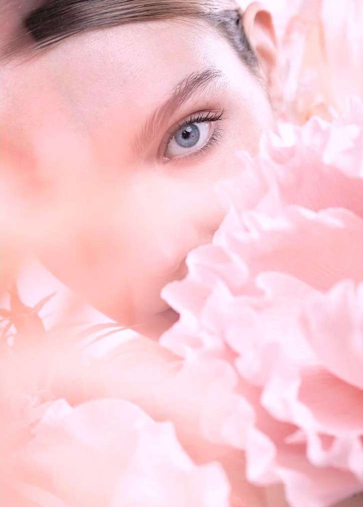Close-up beauty shot of fair-skinned woman's face partially obscured by a mass of soft pink ruffled petals, soft focus, and gentle lighting.