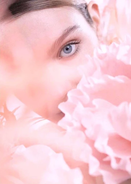 Close-up beauty shot of fair-skinned woman's face partially obscured by a mass of soft pink ruffled petals, soft focus, and gentle lighting.