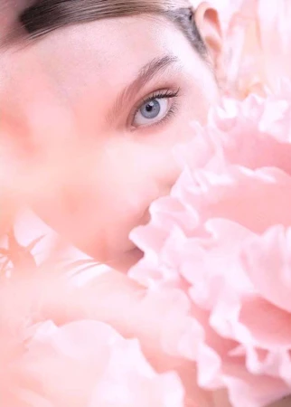 Close-up beauty shot of fair-skinned woman's face partially obscured by a mass of soft pink ruffled petals, soft focus, and gentle lighting.
