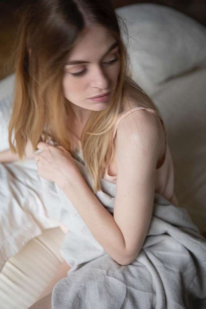 Close-up portrait of a woman with long blonde hair wrapped in a grey blanket on a bed, captured by photographer Sophie Le Gendre.