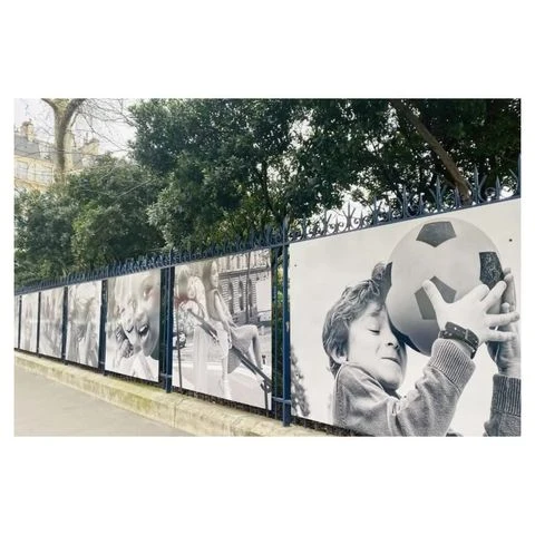 Outdoor art exhibit on a wrought iron fence features black and white photos of children and a soccer ball in an urban environment.