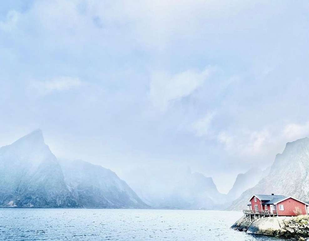 Scenic view of a red house on a rock formation, surrounded by water and mountains under a bright, cloudy sky. Landscape photography.
