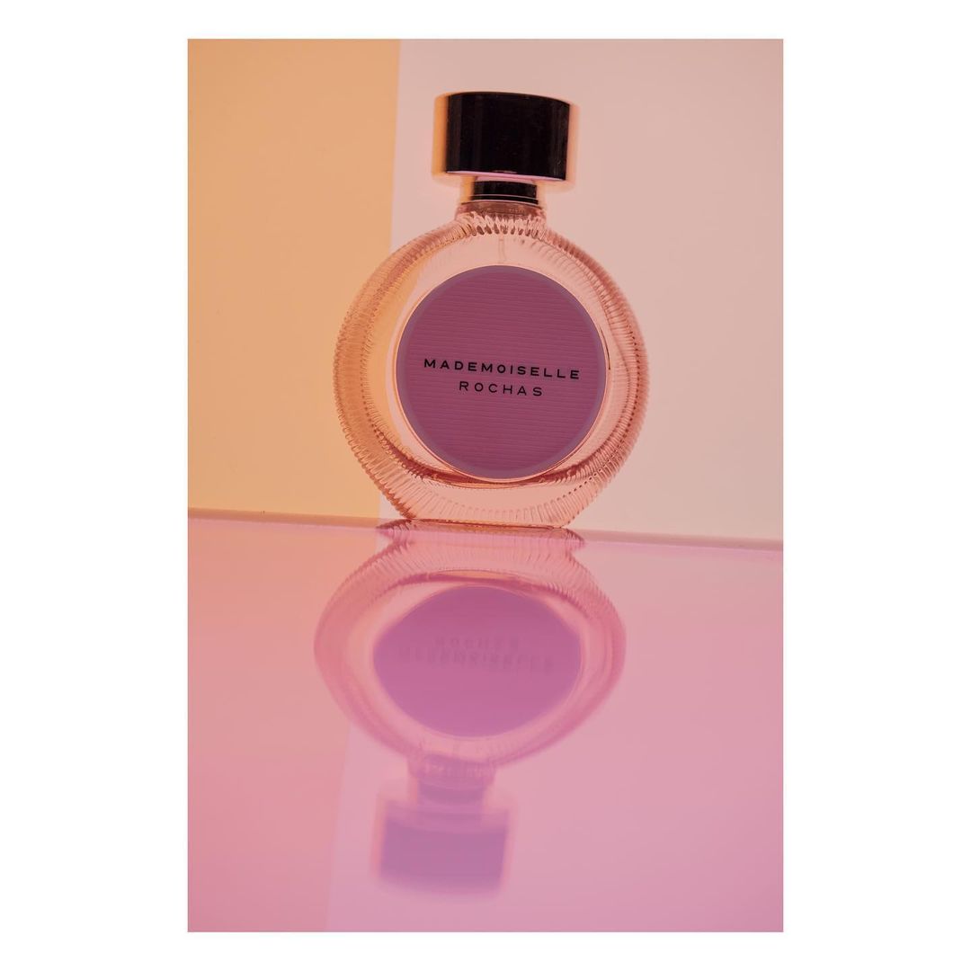 Round Mademoiselle Rochas perfume bottle stands on a reflective pink surface with a peach and white background creating a soft, elegant look.