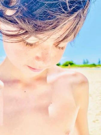 Close-up portrait of a young person with dark hair and freckles outdoors with a sandy beach and bright sky in the background.