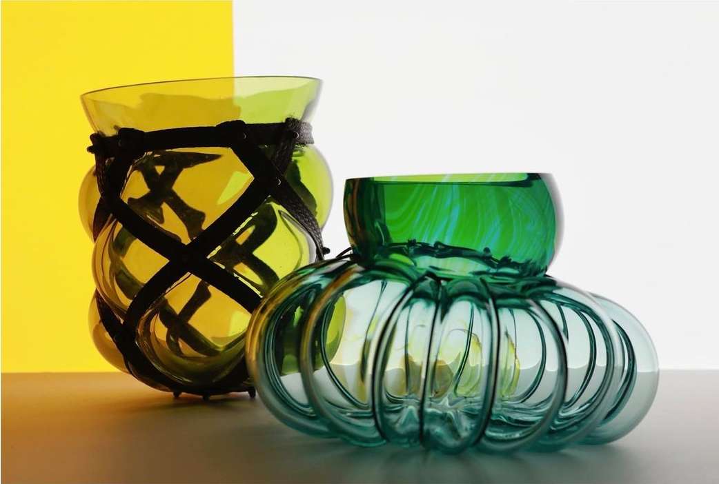 Two glass vases, one green with ridged pattern, the other yellow with black strap detail, against a yellow and white background.