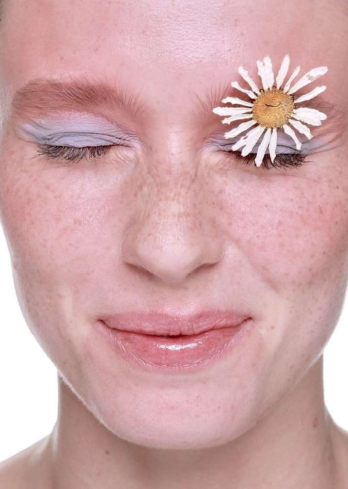 Close-up portrait of a woman with closed eyes and freckles, adorned with a white daisy flower over her eye on a white background.