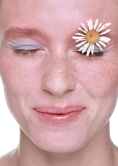 Close-up portrait of a woman with closed eyes and freckles, adorned with a white daisy flower over her eye on a white background.