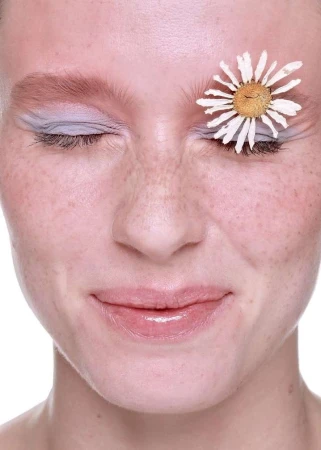 Close-up portrait of a woman with closed eyes and freckles, adorned with a white daisy flower over her eye on a white background.
